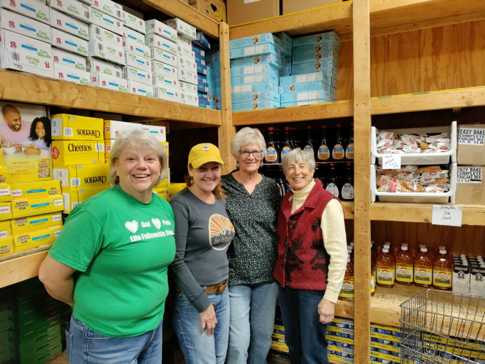 Four smiling volunteers stand in a food pantry, surrounded by shelves stocked with cereal, syrup, juice, and other essentials