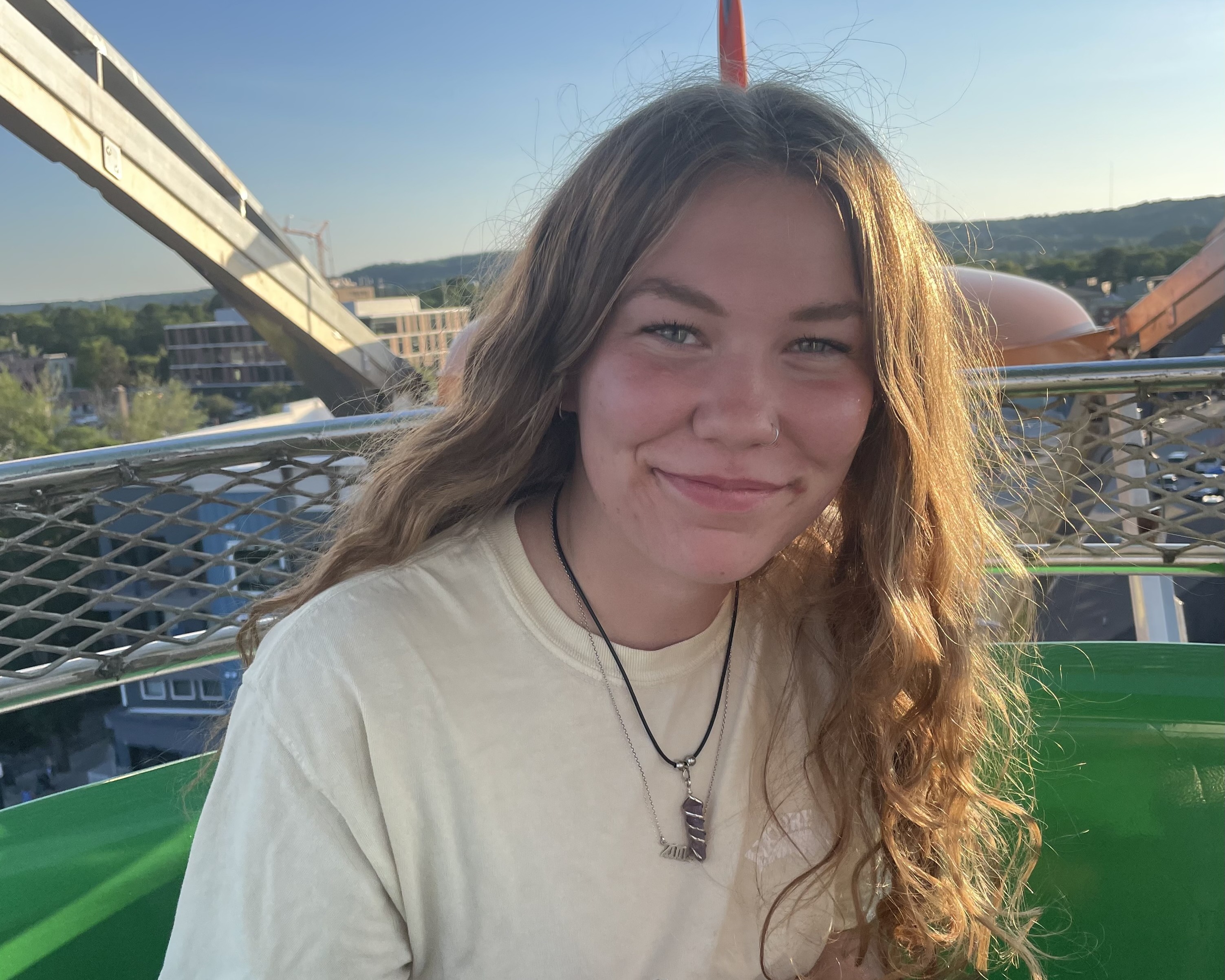 Alana Kuhlen smiles with long hair, wearing a light-colored shirt and necklaces, sits in an amusement park ride. The background shows a clear sky, distant hills, and park structures.