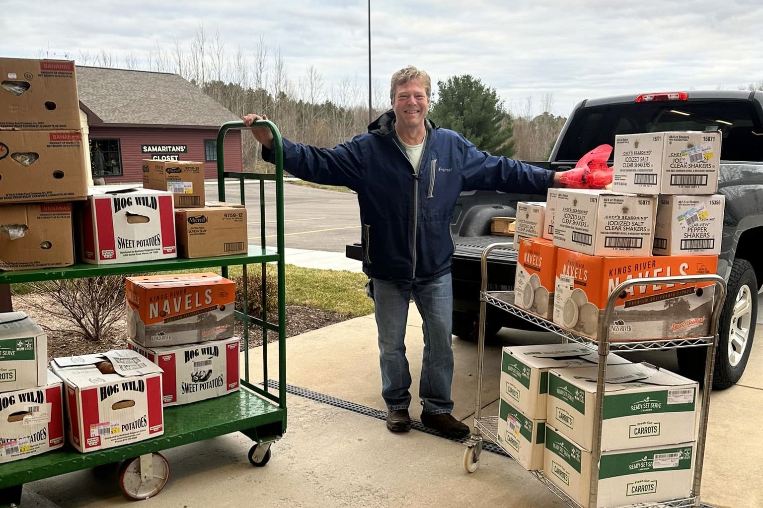 Man standing outdoors between two food carts filled with boxes of produce and supplies, near a building labeled Samaritan’s Closet.