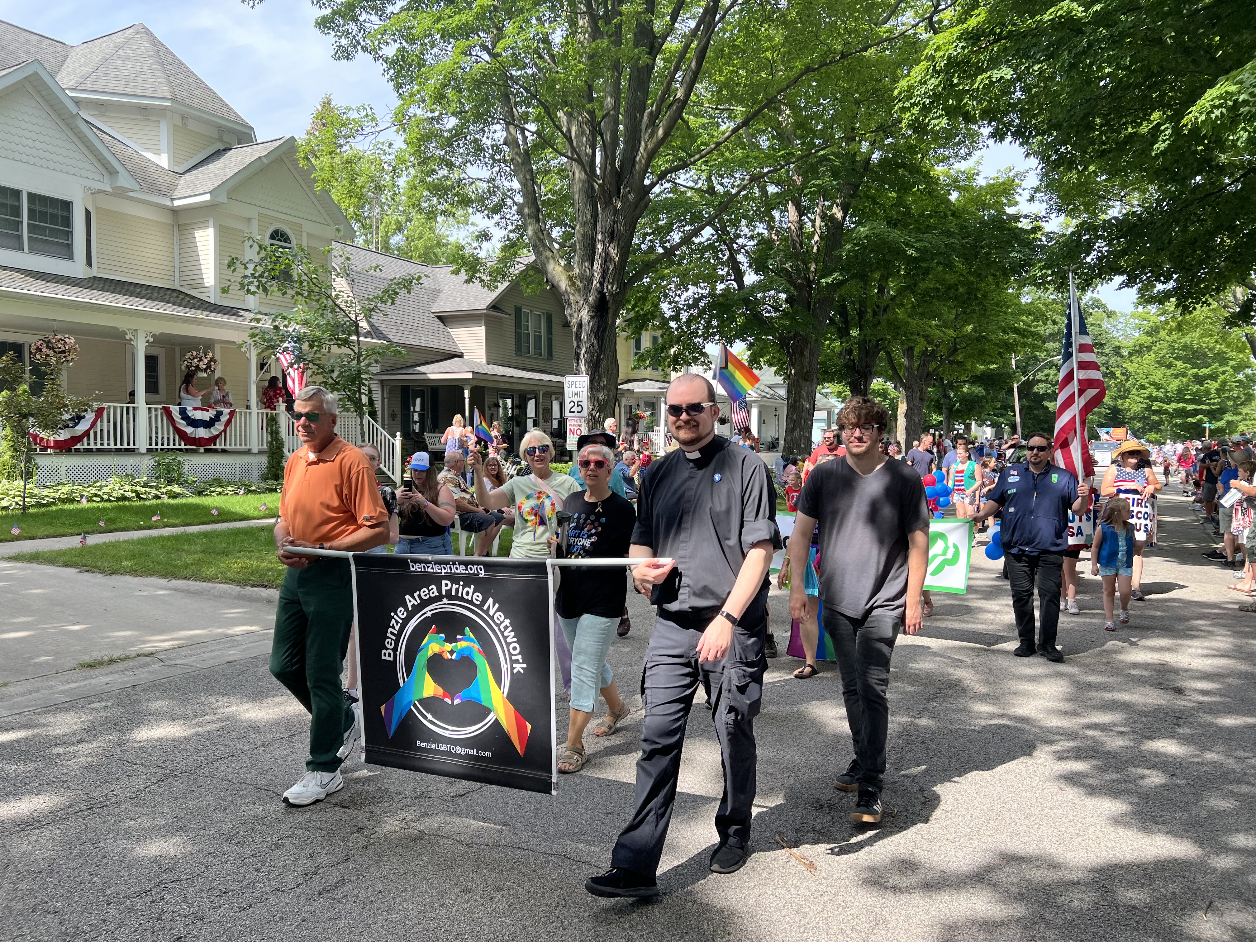 Benzie Pride Network parade with participants holding a banner and Pride flags on a tree-lined street