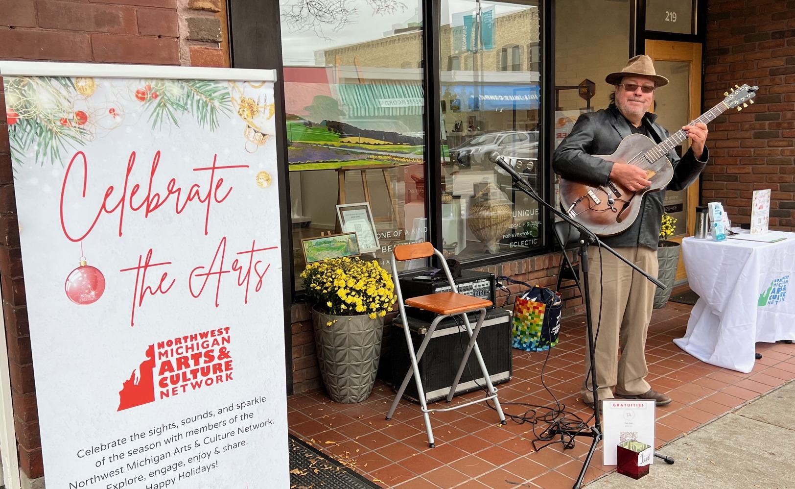 A street performer plays an electric guitar in front of a storefront with a “Celebrate the Arts” sign by the Northwest Michigan Arts & Culture Network, surrounded by yellow flowers and music equipment.