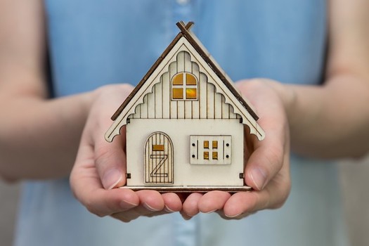 A person holds a small decorative house model in both hands. The light-colored wooden house features detailed windows, a front door, and a pitched roof, symbolizing safety, shelter, and home.
