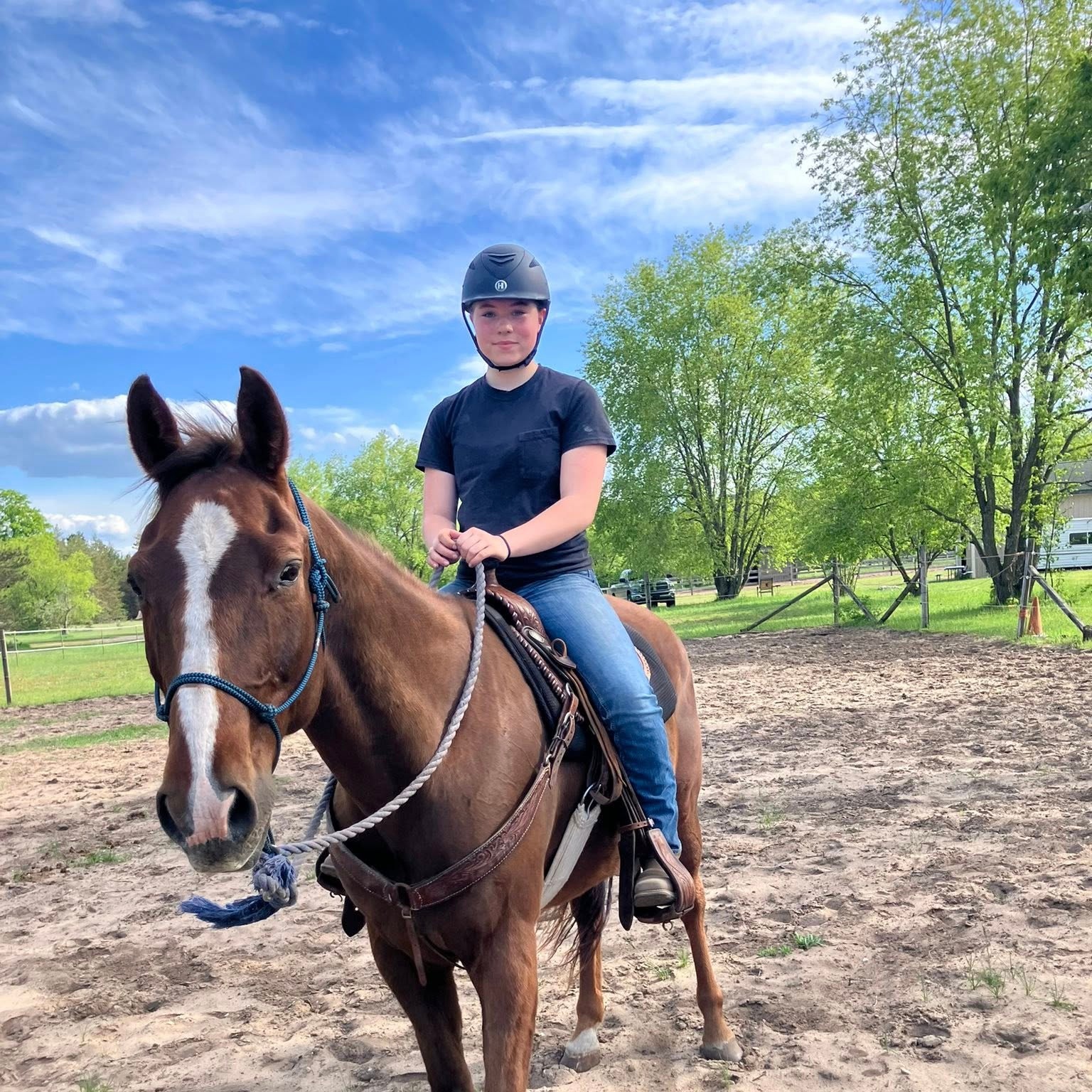 A horseback rider wearing a safety helmet sits on a brown horse in an outdoor riding arena.