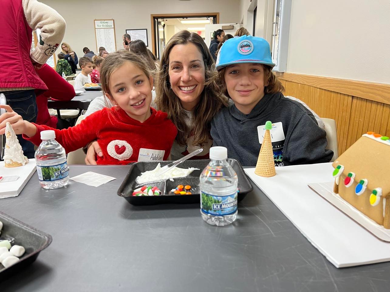 Two children and one adult sit at a table doing a craft activity with decorated gingerbread houses and candy. One child wears a red sweater with a white heart, another wears a blue cap and dark hoodie. Others are participating in the background.