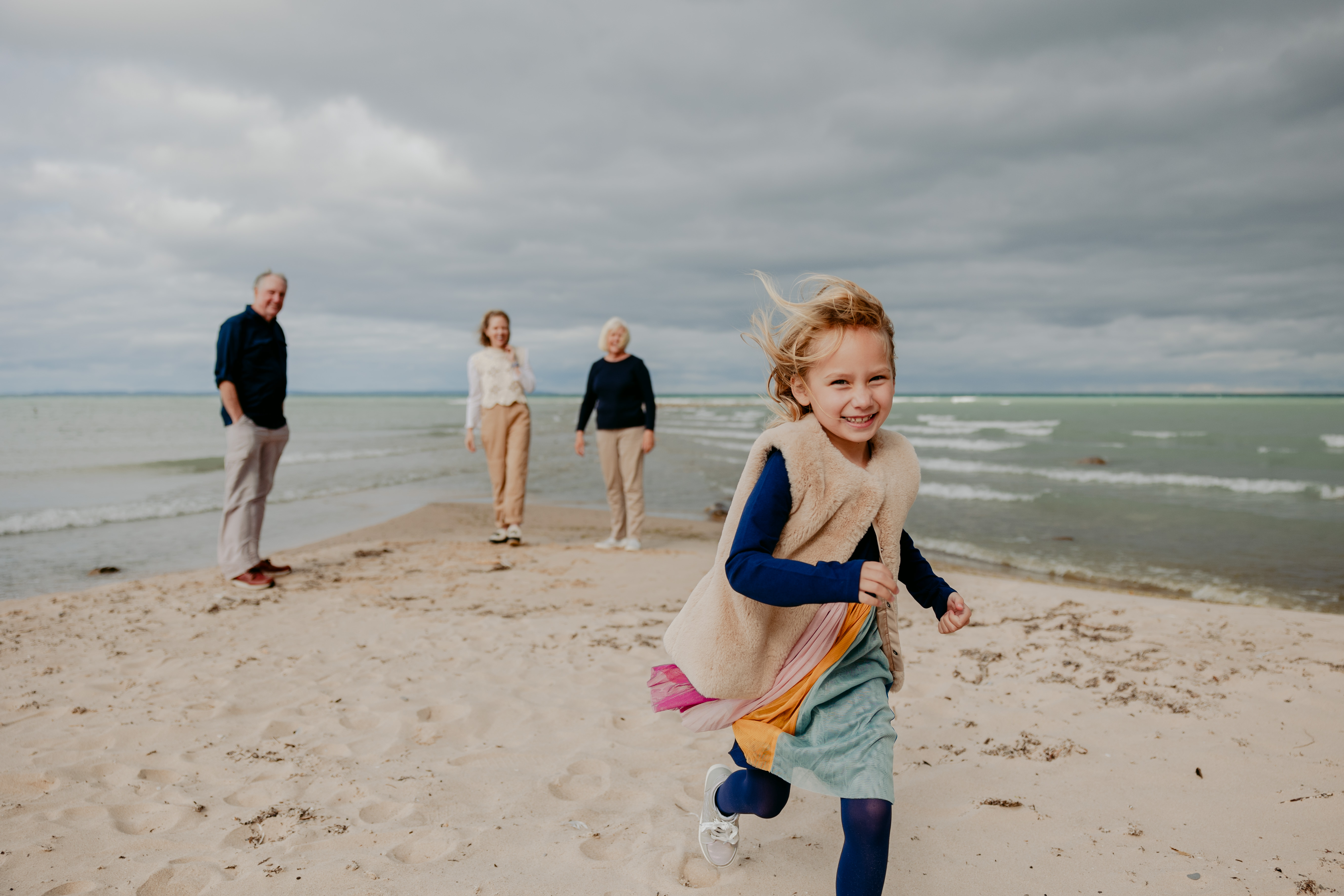 Young girl smiling and running toward the camera on a sandy beach, with waves in the background and three family members—two women and a man—standing and smiling in the distance under a cloudy sky.