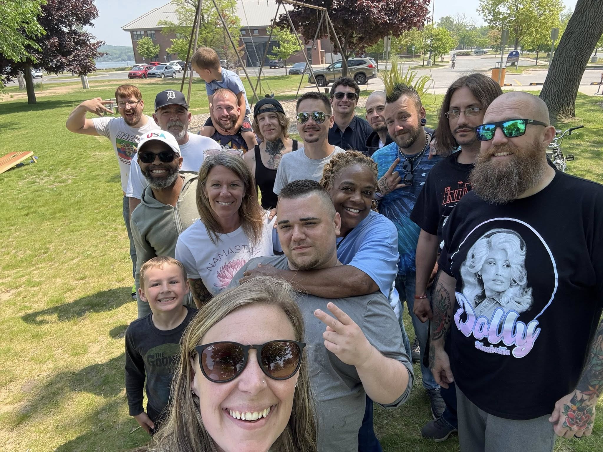 A large group of community members gathers outdoors on a sunny day for a group selfie in a grassy park. Several adults and children pose together near a playground, with trees, parked cars, and a waterfront building visible in the background.