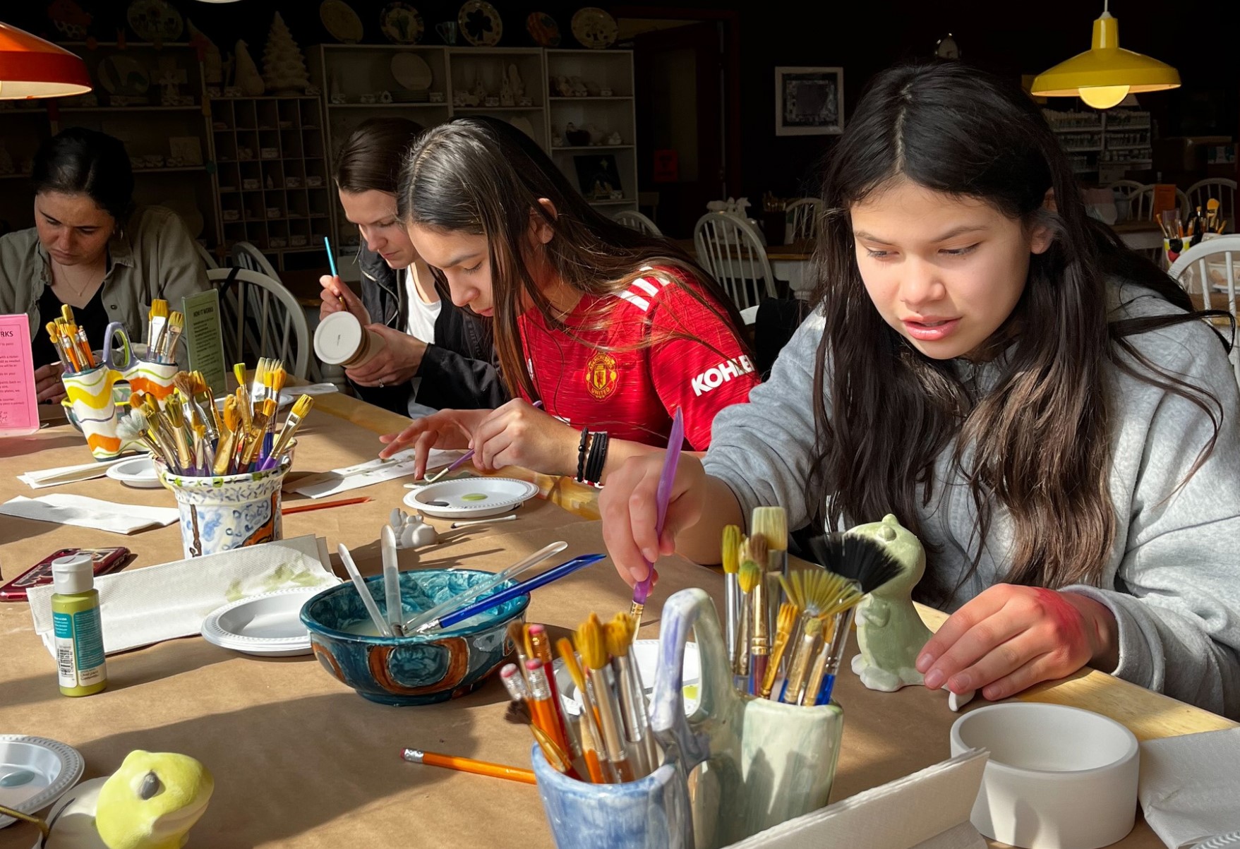 A group of teens sit at a long table in an art studio, painting small pottery pieces. Paintbrushes, palettes, and ceramic supplies are spread across the table in bright sunlight.