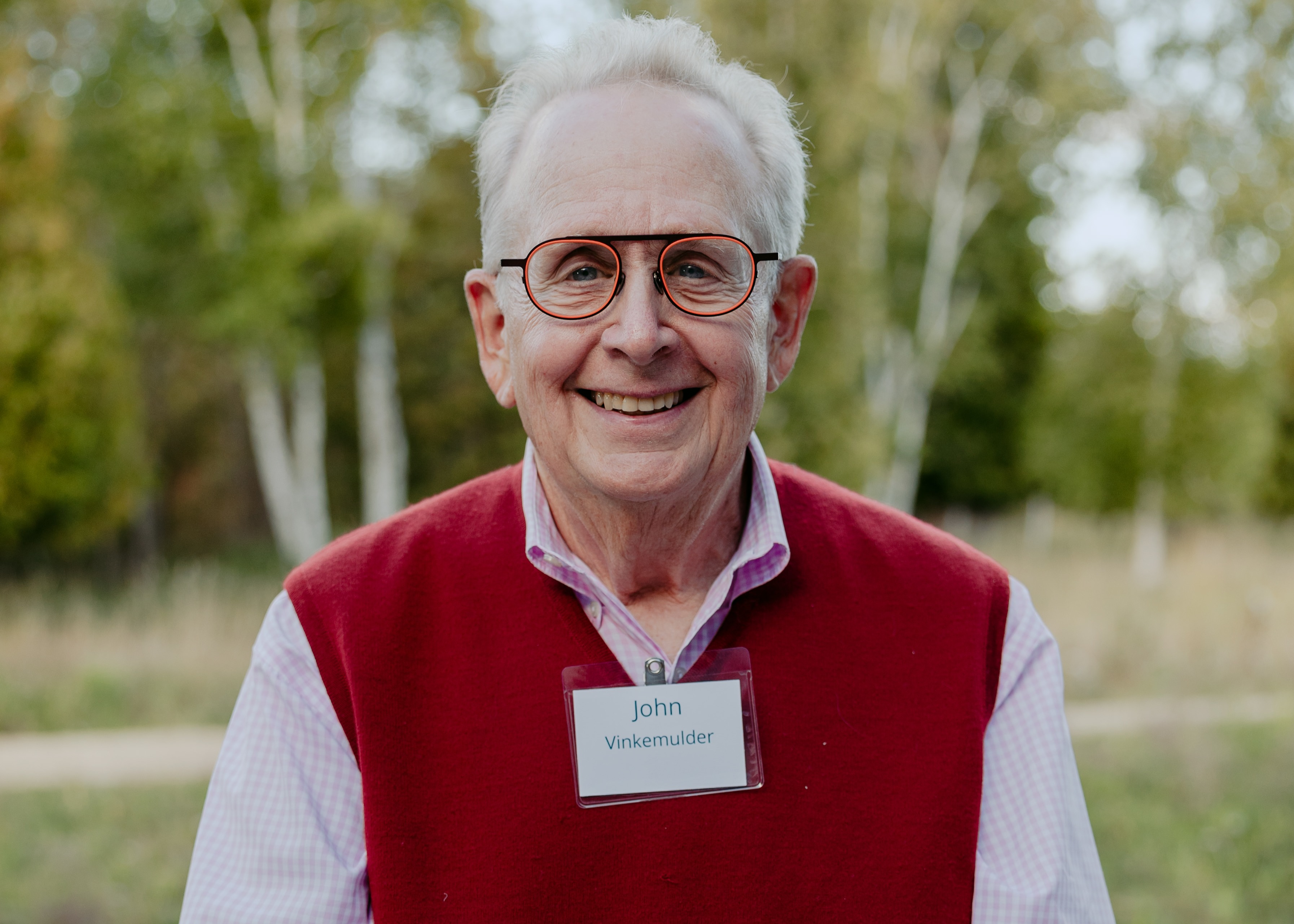 John Vinkemulder (Vink) standing outdoors in a red vest with a name tag, surrounded by trees and greenery.