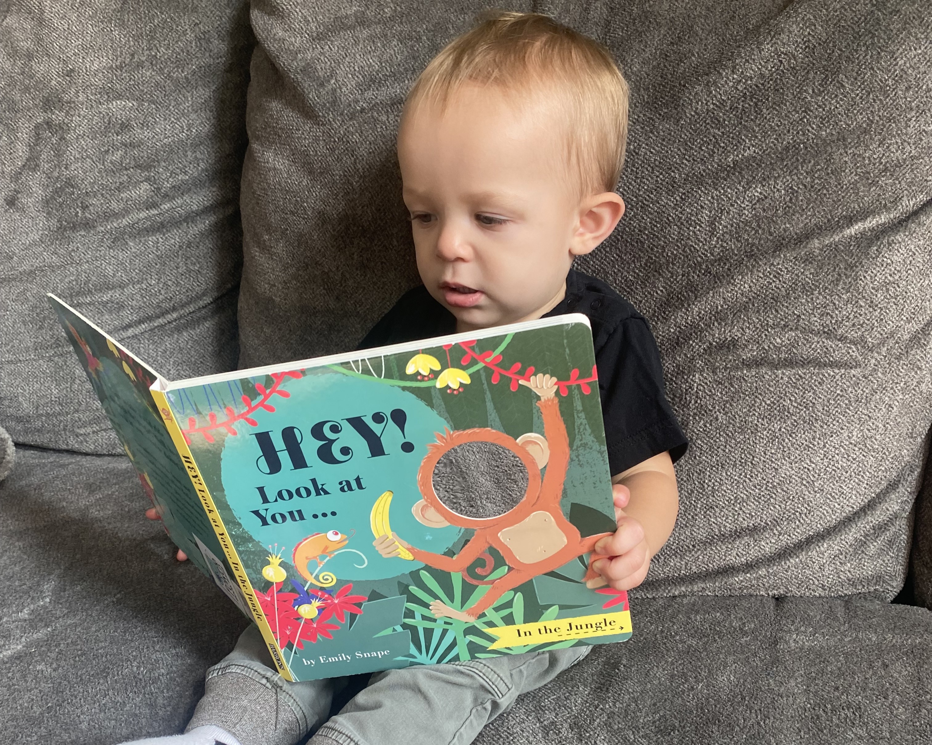 A child is sitting on a gray couch, holding and looking at an open picture book titled “Hey! Look at You!” 