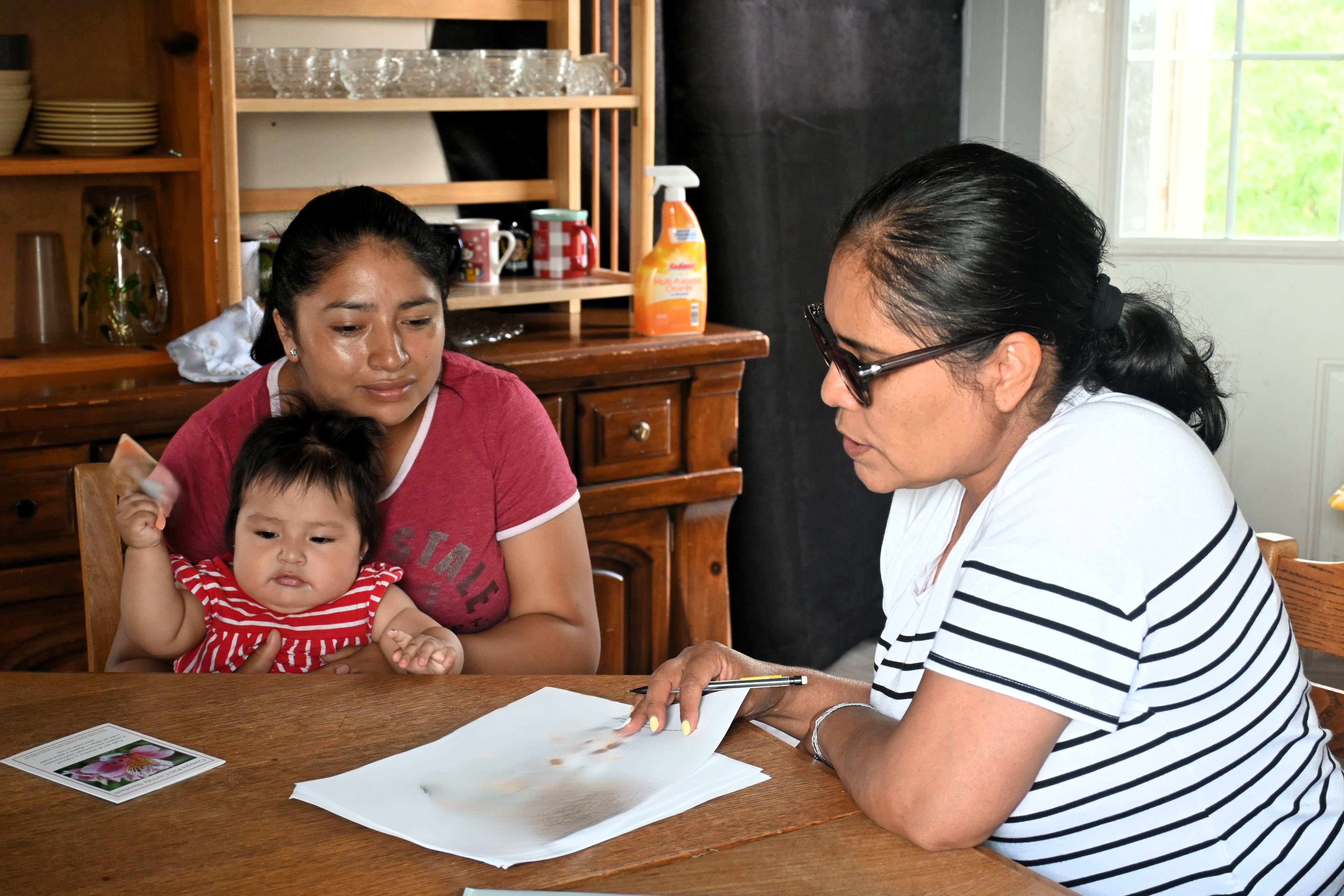 A woman holds a young child on her lap at a wooden table while another woman sits across from them reviewing documents with a pen in hand. 