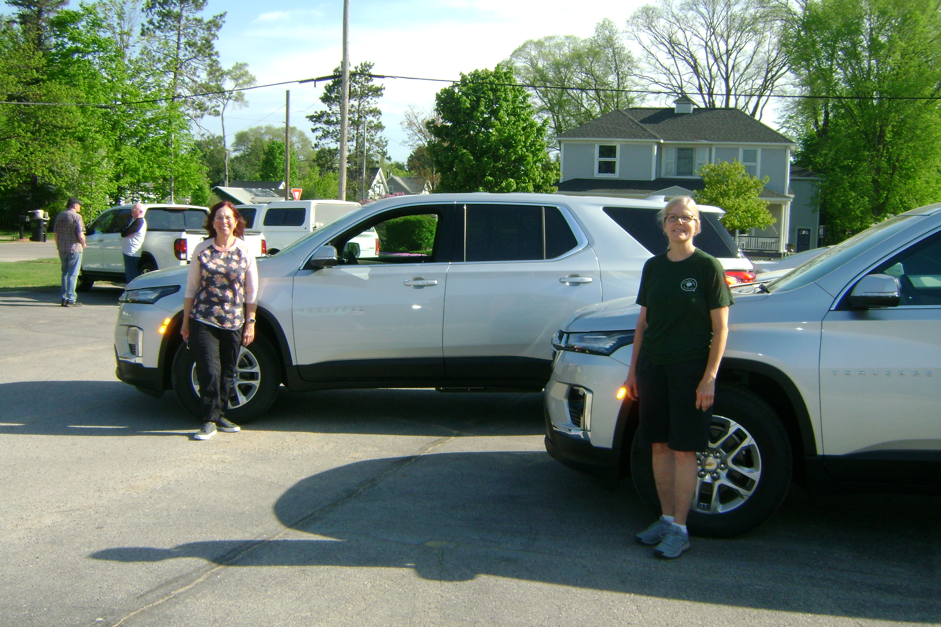 Two Meals on Wheels volunteers stand beside white SUVs in a parking lot on a sunny day.