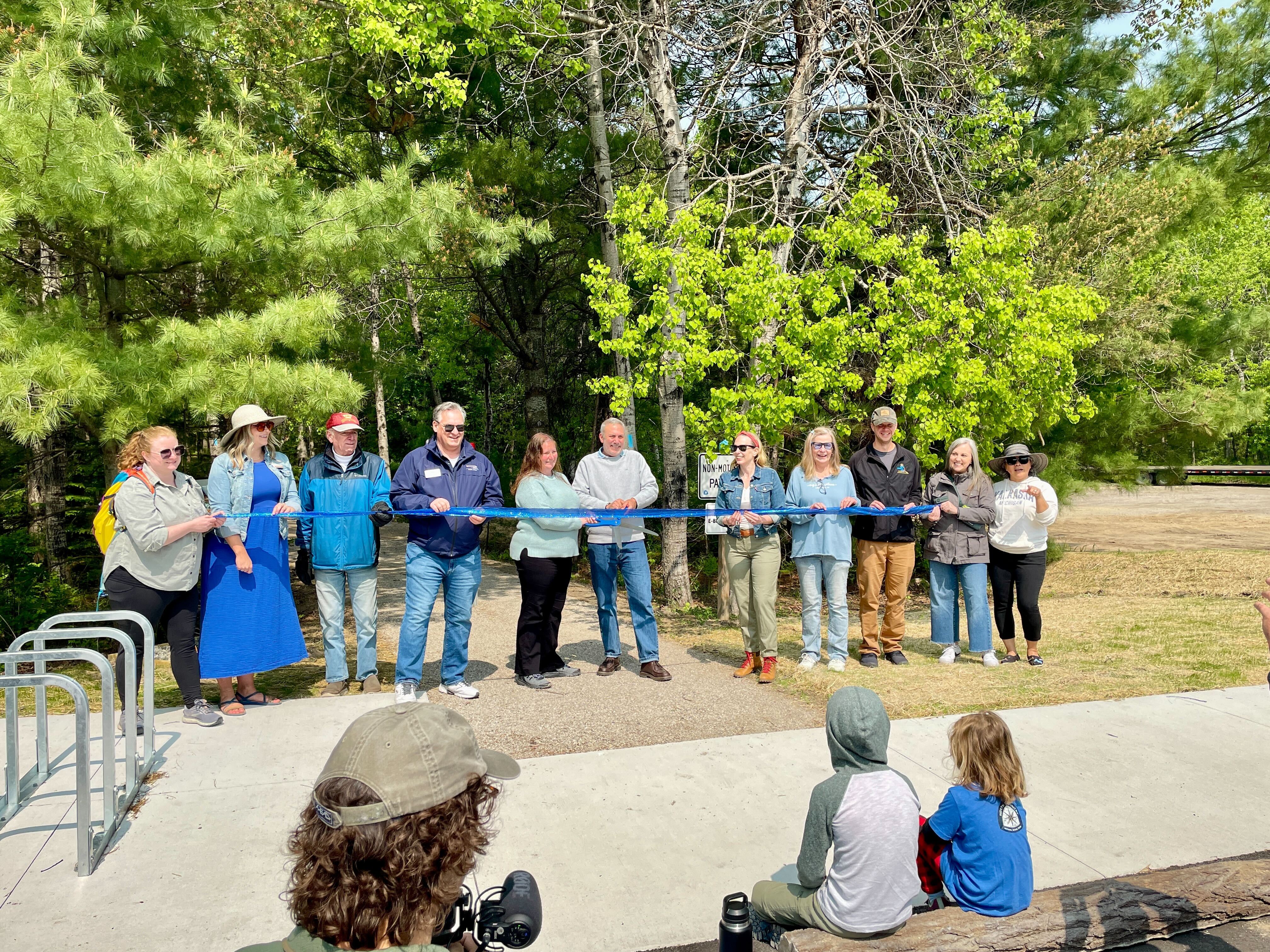 People hold a blue ribbon during an outdoor ribbon-cutting ceremony, with children watching from a bench in a wooded park setting.