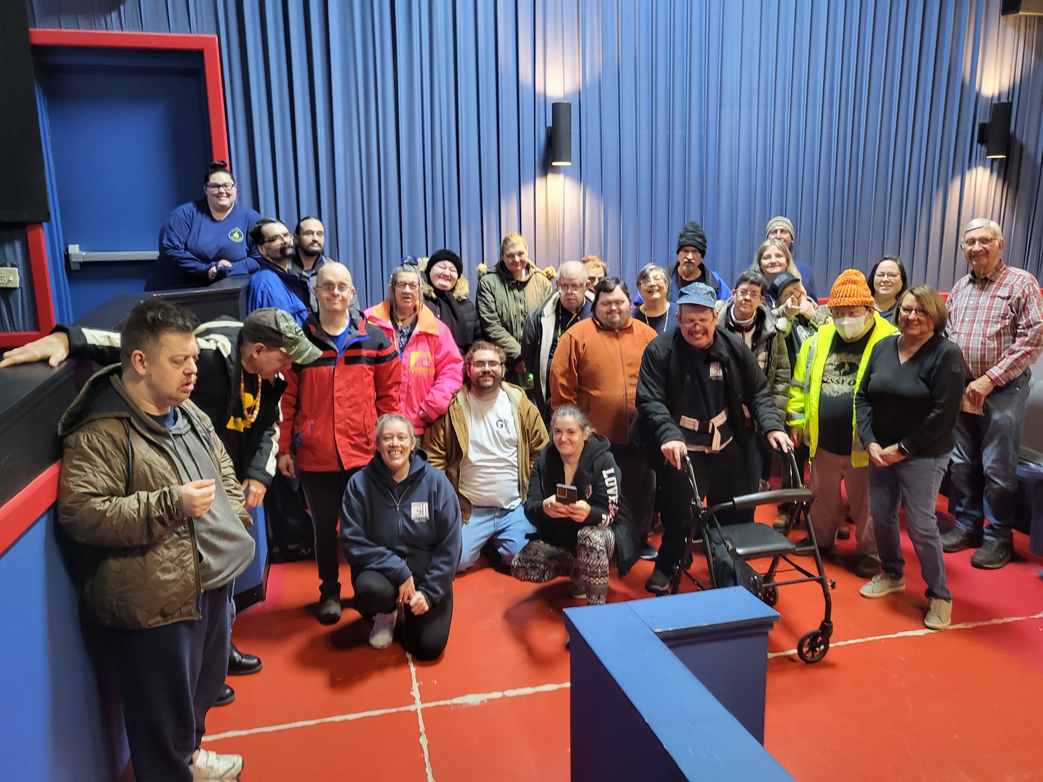 A group of 26 people, some standing and others sitting or kneeling, gathered in a room with blue walls, red flooring, and blue curtains. One person uses a walker