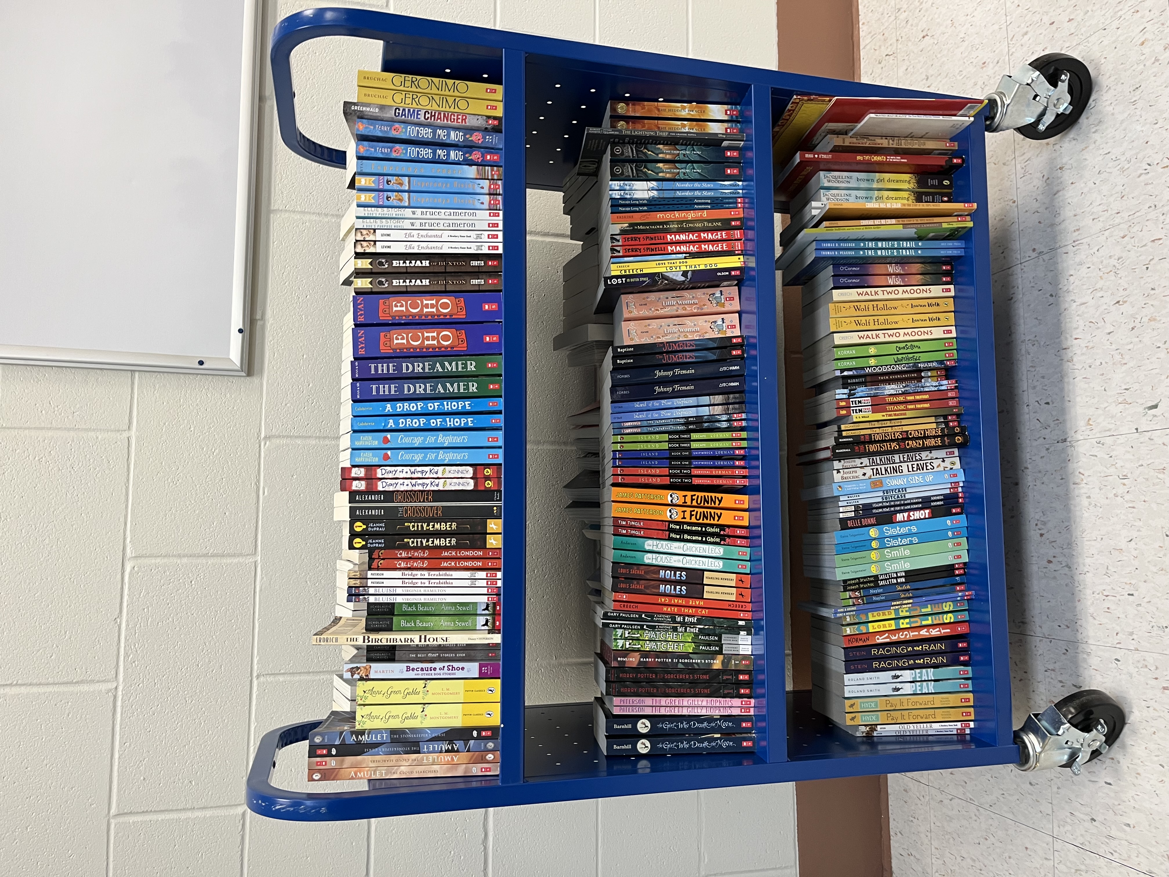 A blue rolling book cart filled with children's and young adult books in a school or library setting.