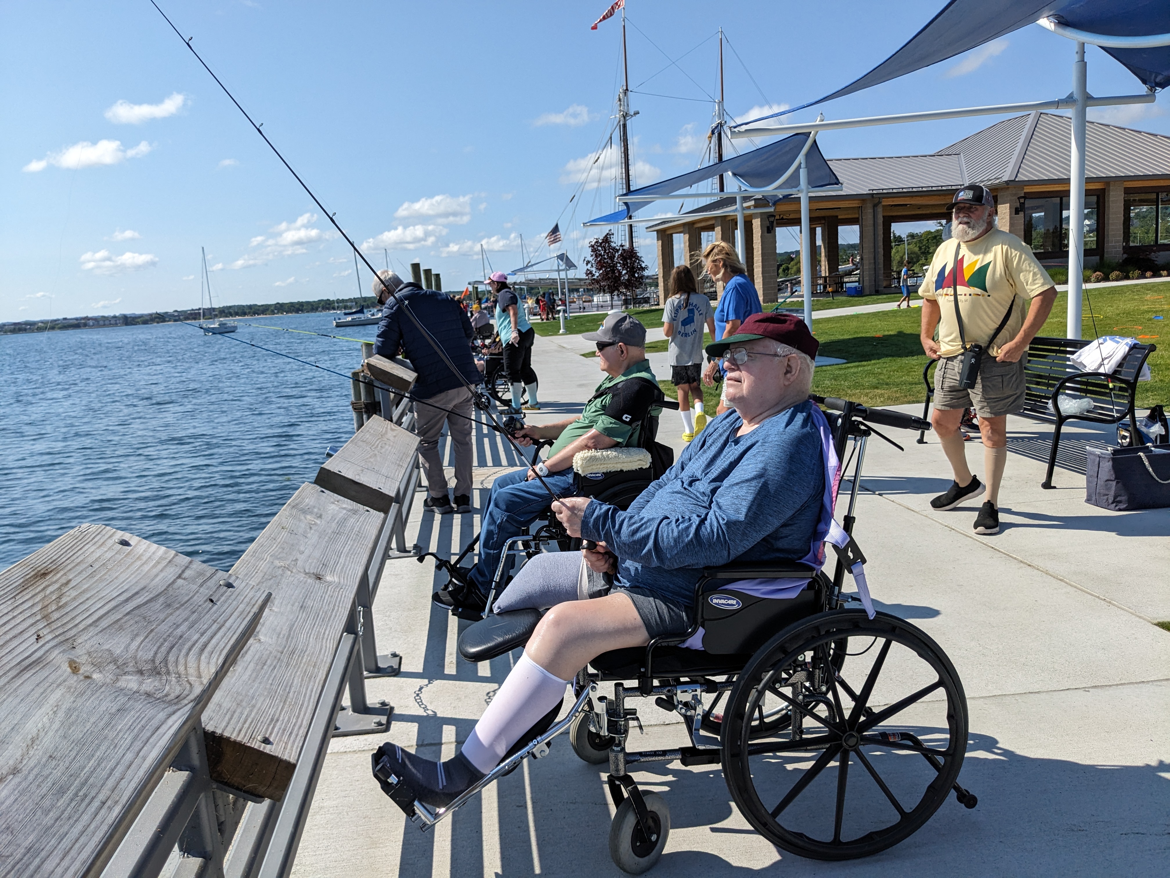 Individuals fishing from a pier beside calm water, with wheelchairs, benches, and shade structures visible, and boats in the distance.
