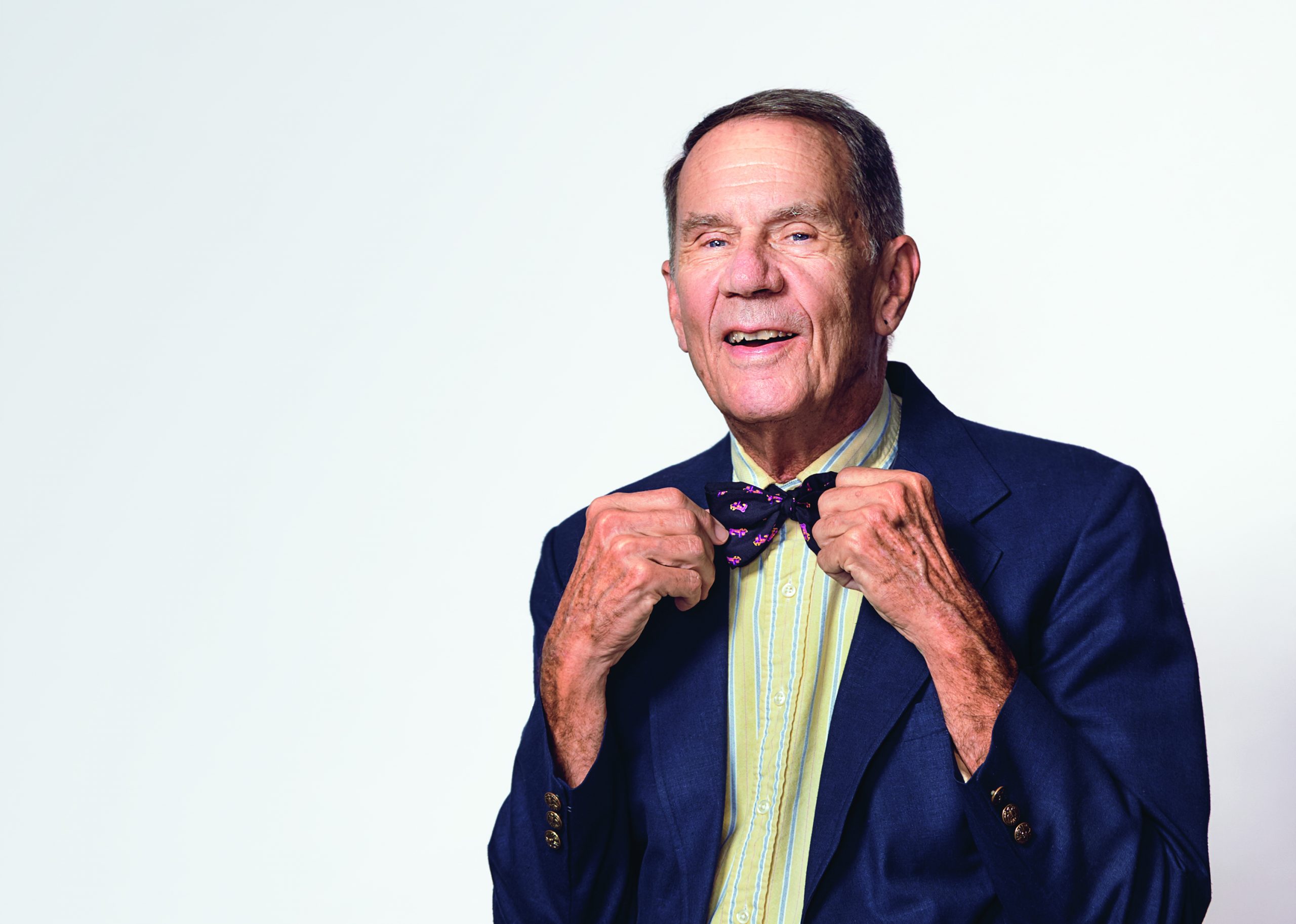 Micheal Dively, smiling, adjusting a patterned bow tie while wearing a dark blue blazer over a light green and yellow striped shirt, standing against a plain light grey background.