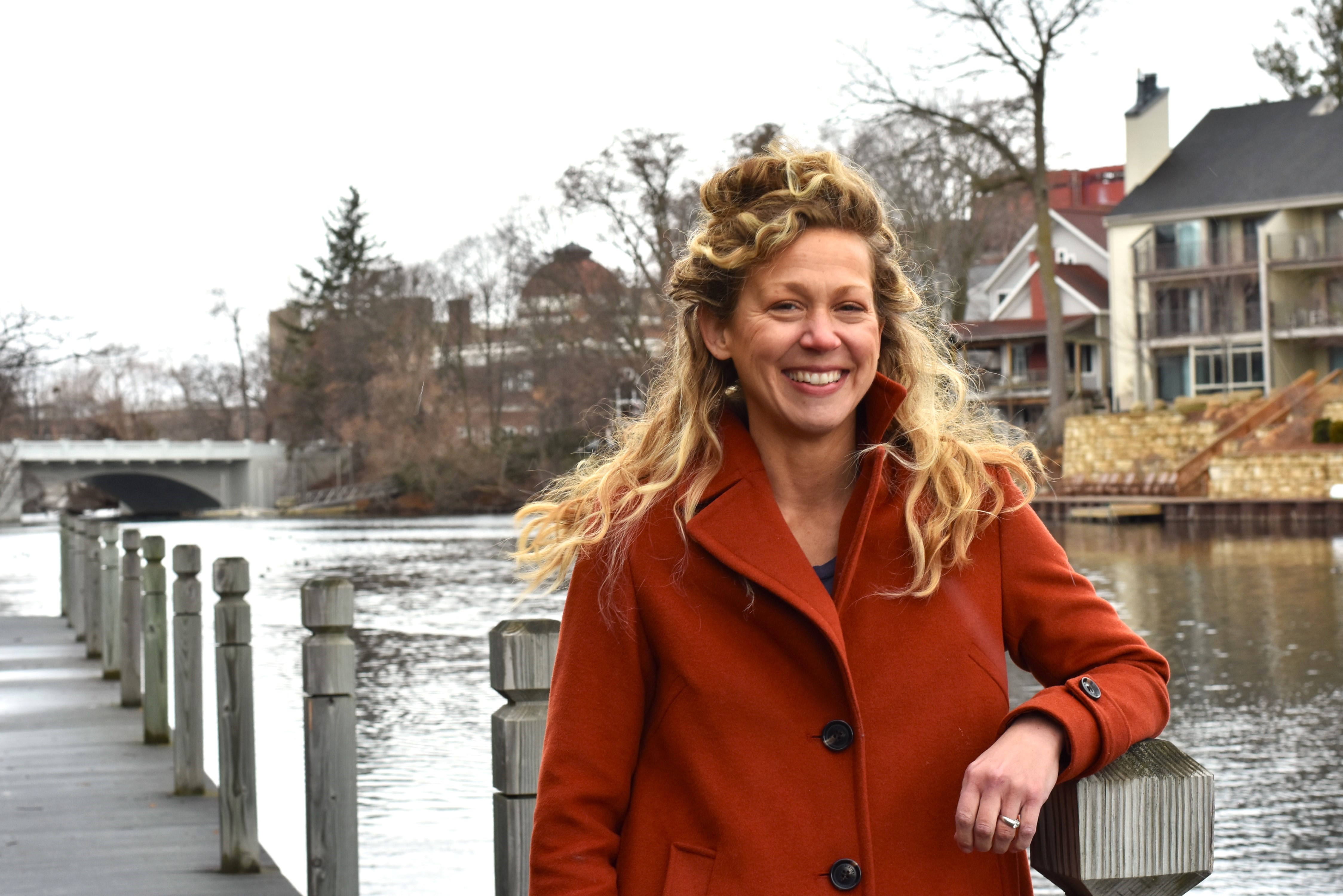 Leah McCallum wears an orange coat with long blonde hair stands on a boardwalk by a river, with trees, houses, and a bridge in the background.