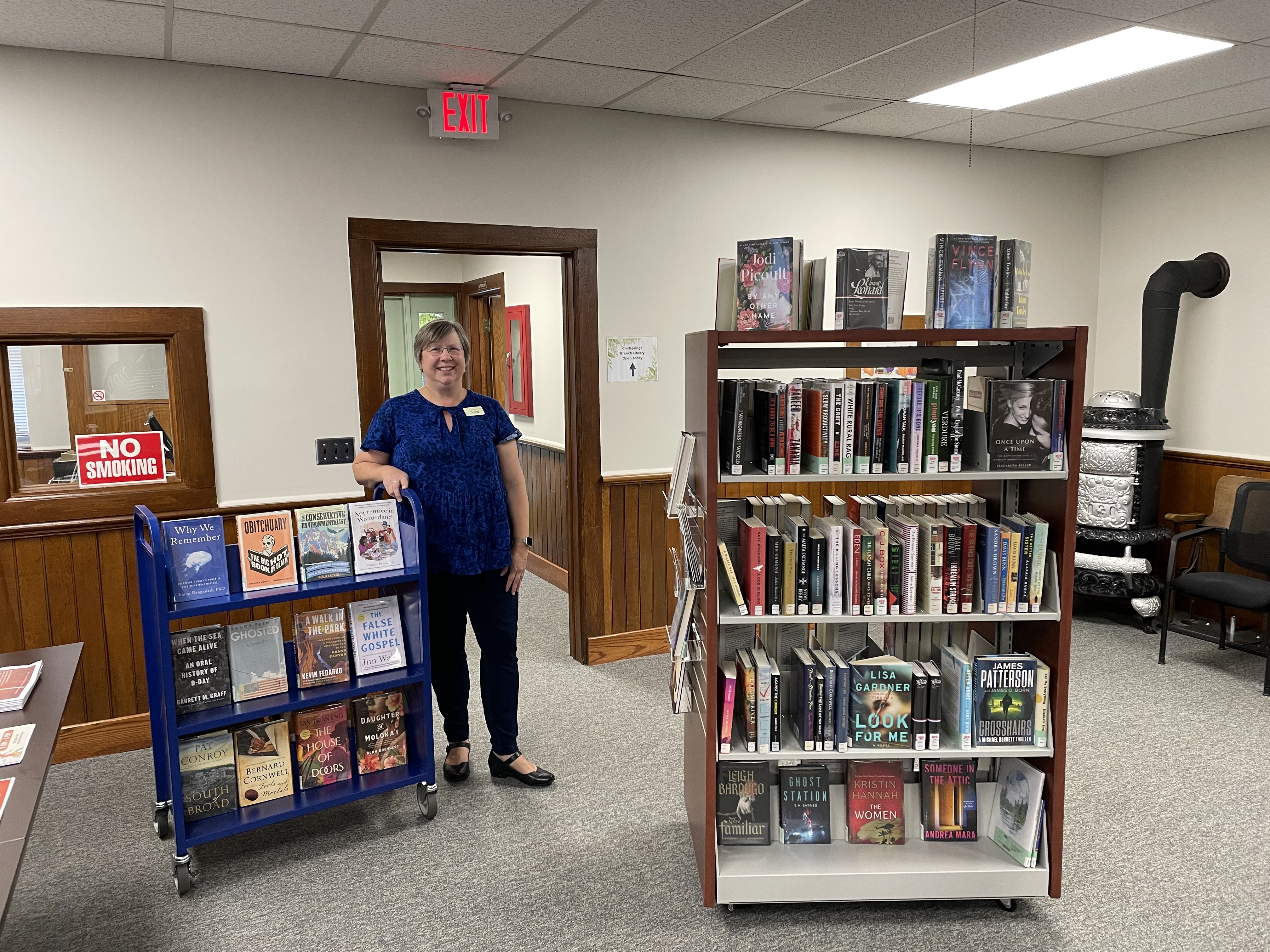 Person wearing blue top and black pants stands, smiling beside a blue book cart and bookshelves full of books at the Kalkaska County Library.