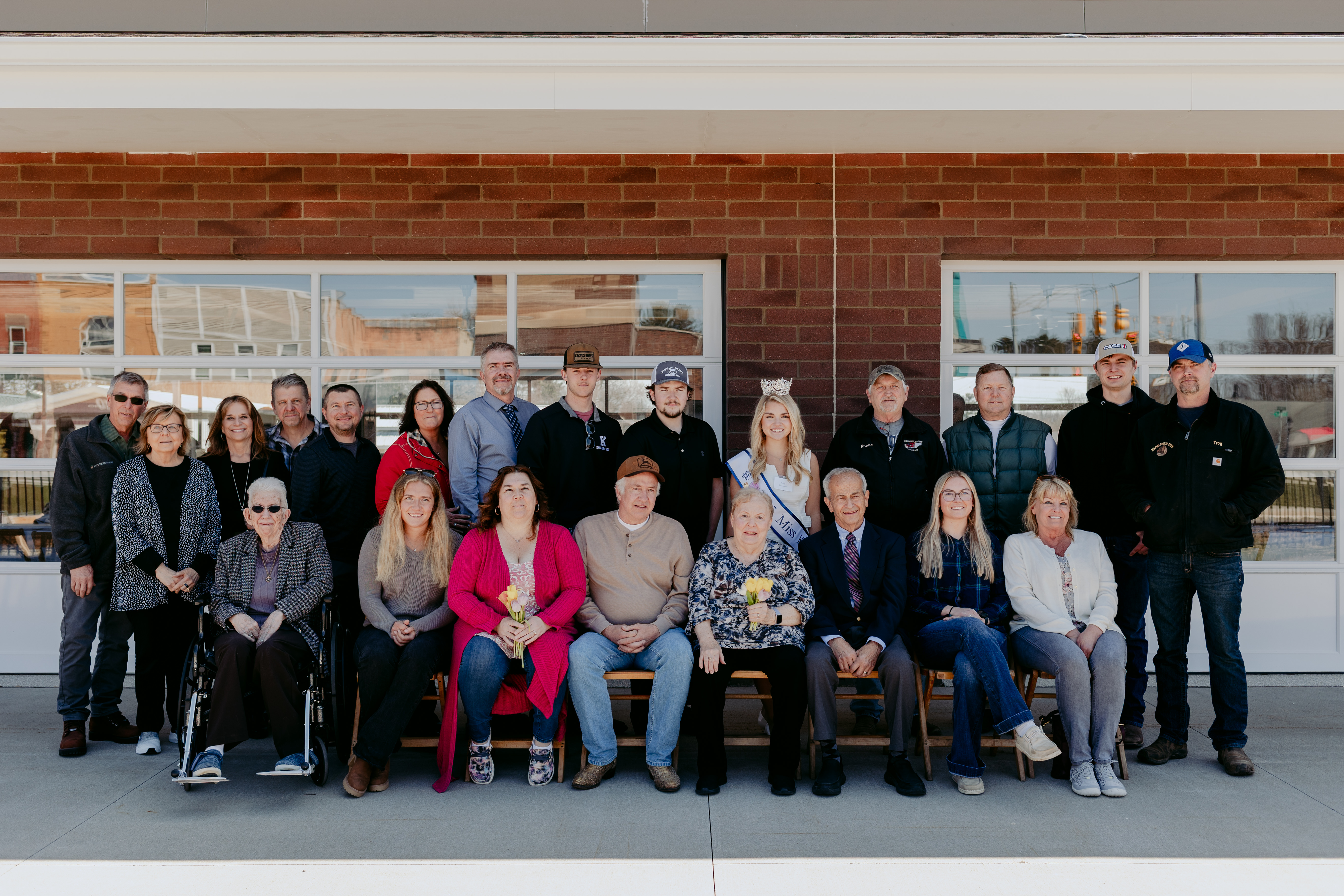 Group photo of 18 people posed in two rows outside a brick building with large windows.