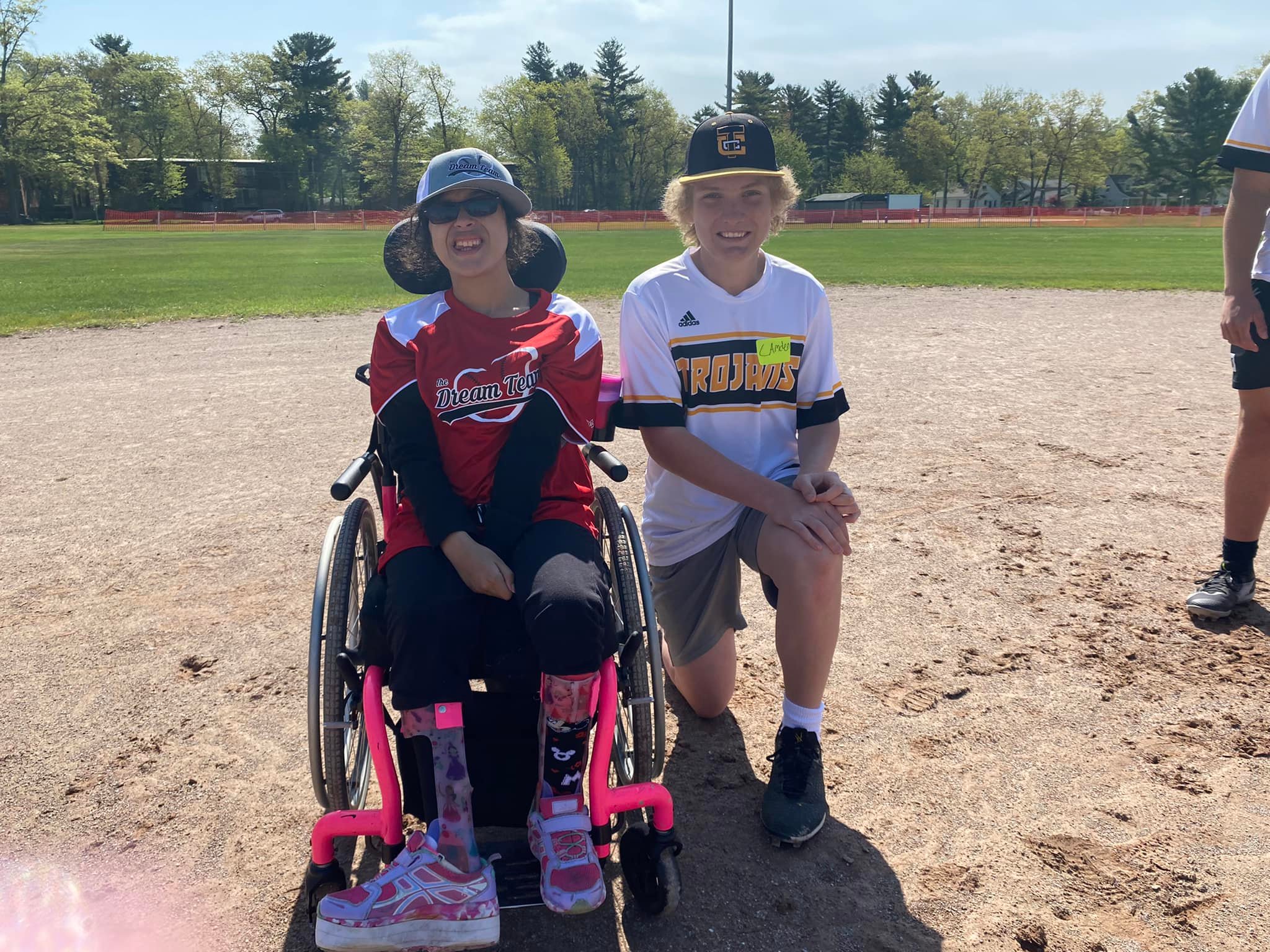 Two people on a baseball field; one in a pink wheelchair wearing a red "Dream Team" shirt, the other kneeling beside them in a white "Trojans" jersey. Trees and grass in the background.