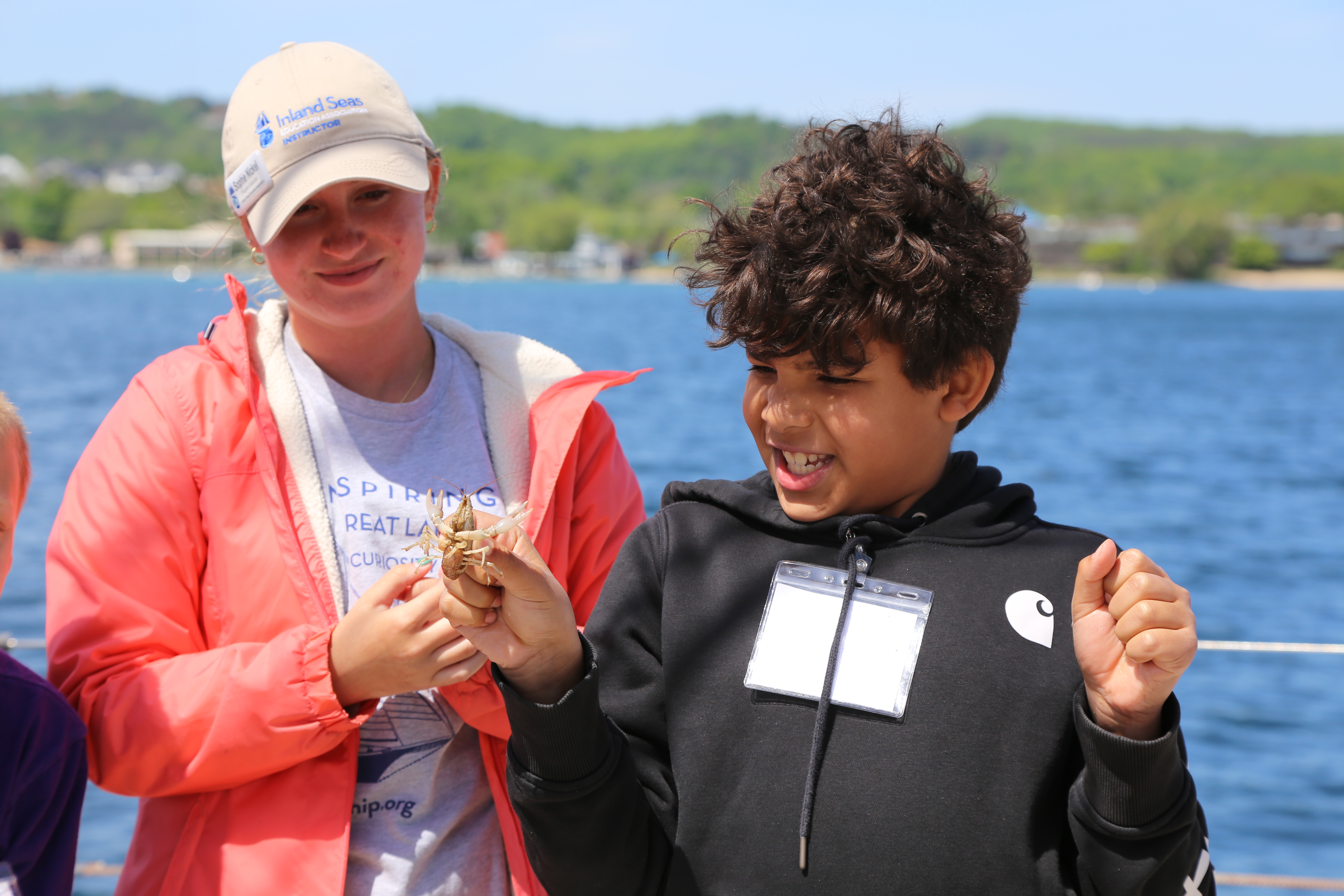 A young boy excitedly holds up a crayfish while smiling, with a woman standing beside him. The woman is wearing a coral jacket, a beige cap, and a t-shirt that says "Inland Seas" and "Great Lakes." The scene is set outdoors on a sunny day, with a body of water and greenery in the background.