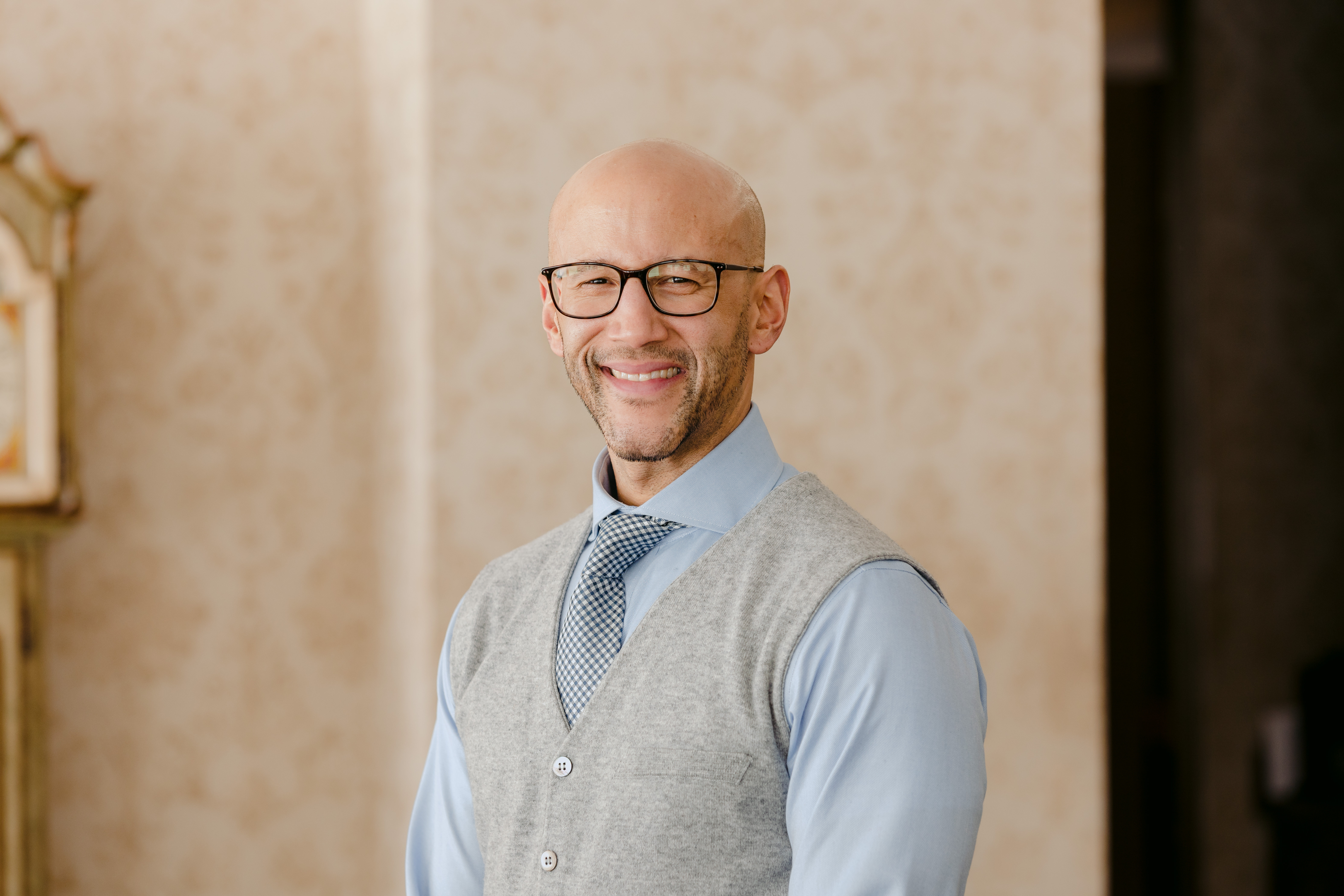 Damian Lockhart wearing a light blue dress shirt, patterned tie, and a gray buttoned vest stands indoors against a softly patterned beige background.