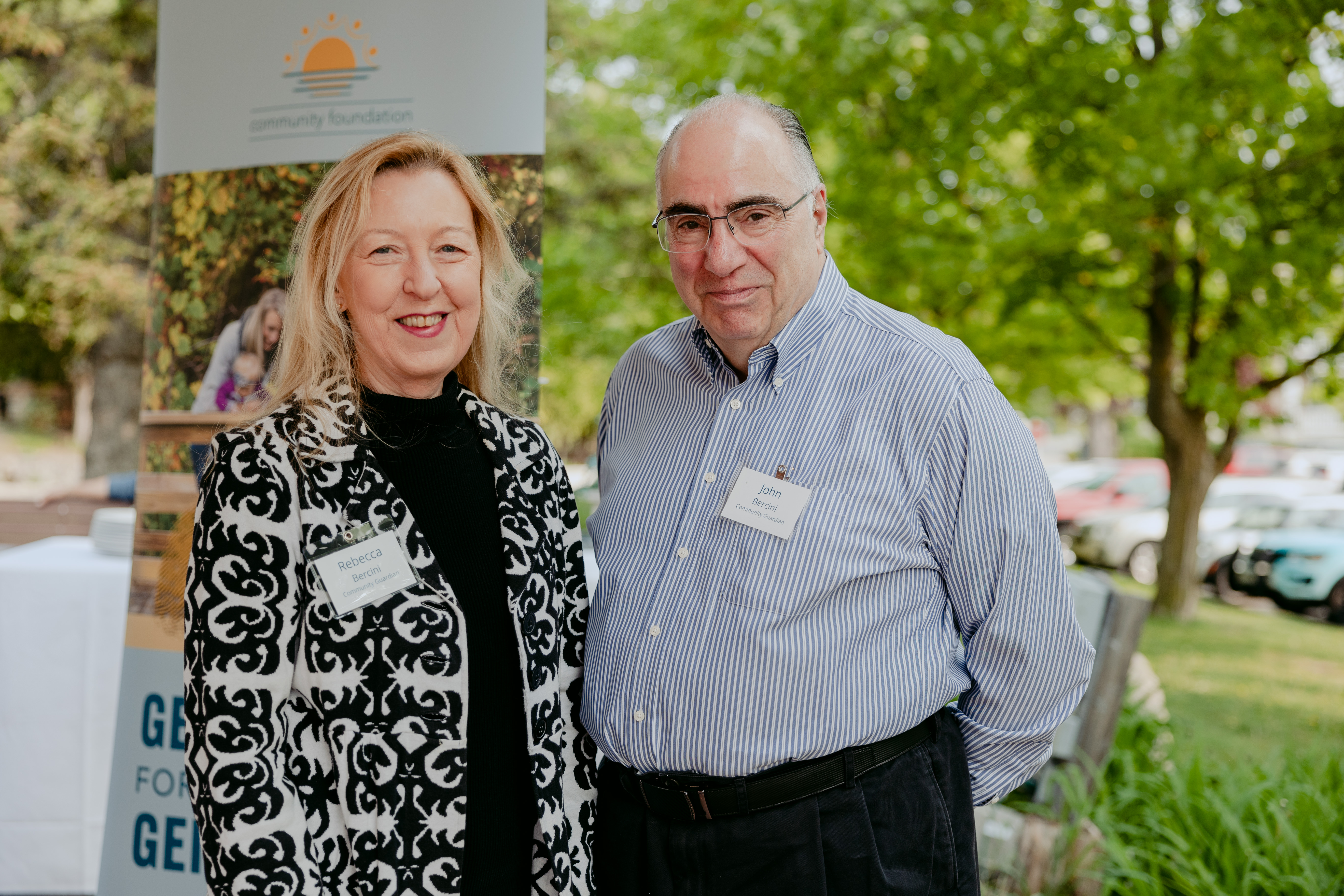 Rebecca and John Bercini smile for a photo. Rebecca wears a black and white blazer and John sports a blue and white striped button down .A banner and greenery is in the background.