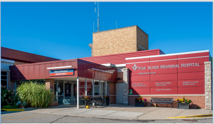 Exterior view of Paul Oliver Memorial Hospital showing the main entrance and emergency signage under a clear blue sky