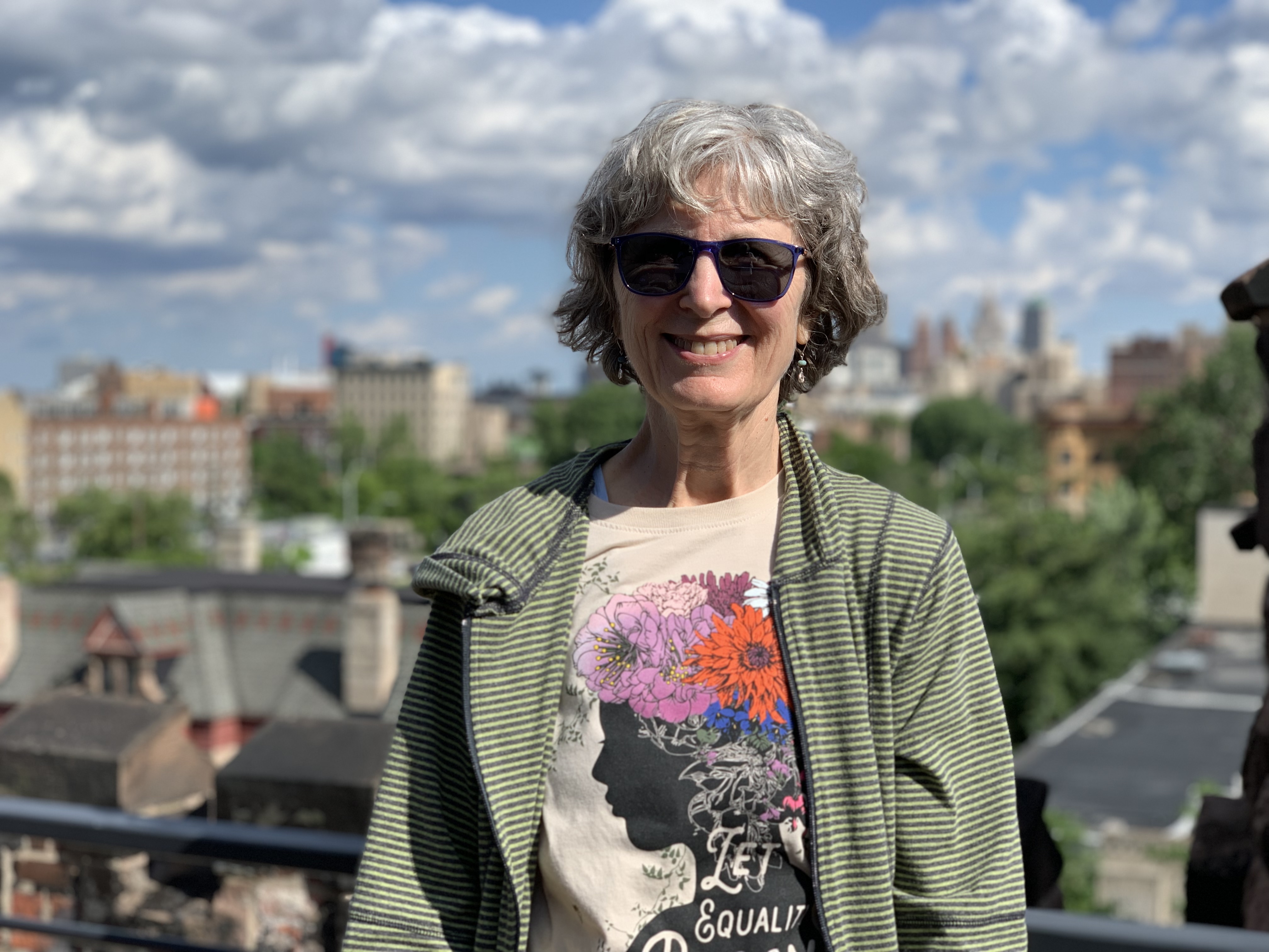 Kathy Tuckerman with short gray hair stands outdoors wearing a striped jacket and a T‑shirt featuring a floral design and silhouette artwork. A cityscape with trees, rooftops, and tall buildings appears in the background under a partly cloudy sky.