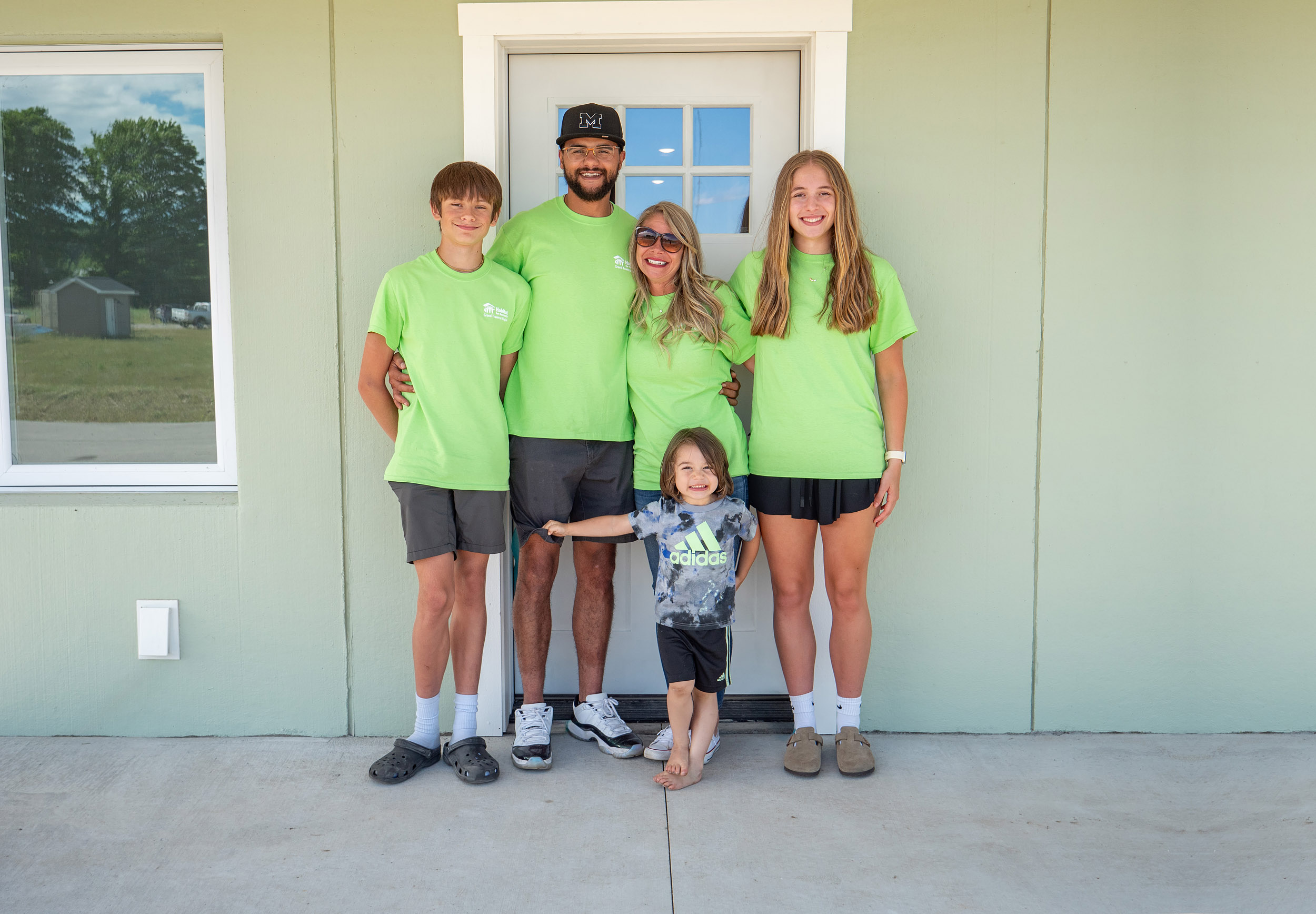 Five people—two adults and three children—stand in front of a light green building, with the adults and two children wearing matching bright green t-shirts and the youngest is in a black Adidas shirt.